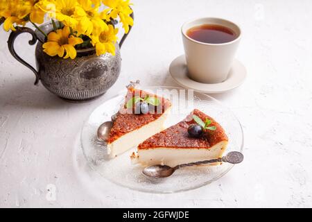 Tranches de cheesecake basque maison brûlé sur plaque avec bleuets et feuilles de menthe, tasse de thé noir sur fond clair avec fleurs dans vase Banque D'Images