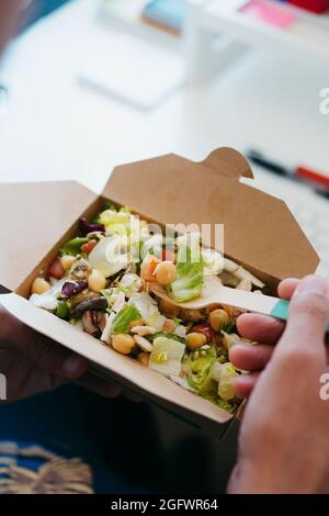 gros plan d'un jeune caucasien assis à son bureau, mangeant une salade de pois chiches dans un contenant en papier, à l'aide d'une fourchette en bambou Banque D'Images