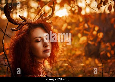 Femme avec des cornes de cerf dans la forêt d'automne. Belle créature gothique mystère dans un long manteau rouge avec des cornes de cerf dans la forêt d'automne. Elf de créature de fantaisie sombre dans le bois magique Banque D'Images