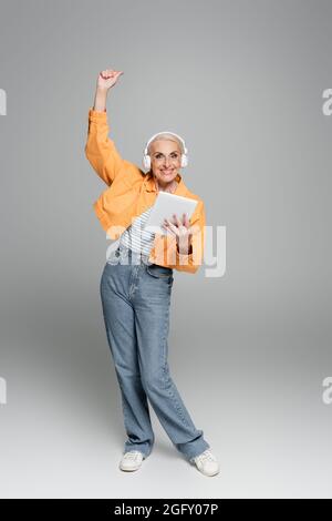 femme âgée joyeuse dans un casque tenant une tablette numérique et montrant le geste de gain sur le gris Banque D'Images