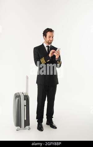 Jeune pilote utilisant un smartphone près d'une valise sur fond blanc Banque D'Images