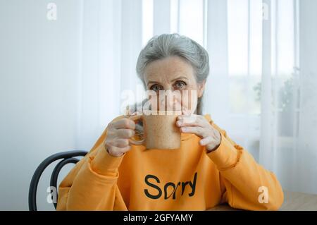 Une femme âgée à cheveux gris boit du café tout en étant assise à la maison. Bonne retraite Banque D'Images