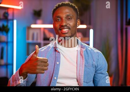 Portrait d'un homme afro-américain souriant dans une chemise en denim montrant le pouce vers le haut tout en posant dans le salon avec la lumière du soir. Des lumières de nuit colorées en arrière-plan. Concentrez-vous sur le visage. Banque D'Images