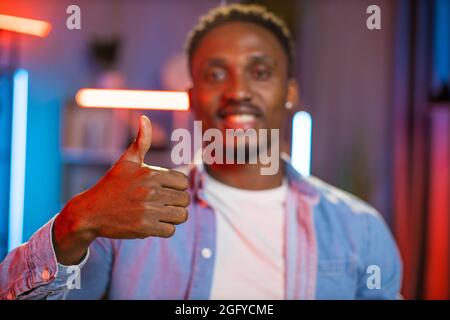 Portrait d'un homme afro-américain souriant dans une chemise en denim montrant le pouce vers le haut tout en posant dans le salon avec la lumière du soir. Arrière-plan flou. Se concentrer sur les doigts. Banque D'Images