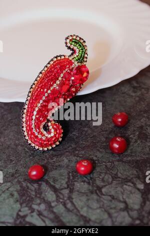 Une broche ornée de perles en forme de poivron rouge bulgare se trouve sur une table en marbre vert devant une assiette blanche vide. Régime alimentaire. Banque D'Images