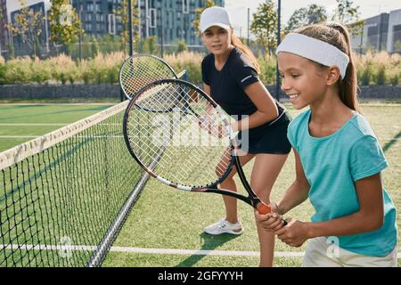 Une petite fille positive tenant une raquette tout en apprenant à jouer au tennis avec son entraîneur féminin sur un court extérieur. Instructeur individuel à un enfant pour téni Banque D'Images