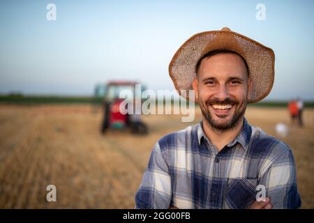 Portrait d'un beau fermier satisfait avec chapeau de paille sur la tête dans le champ pendant la récolte avec tracteur en arrière-plan Banque D'Images