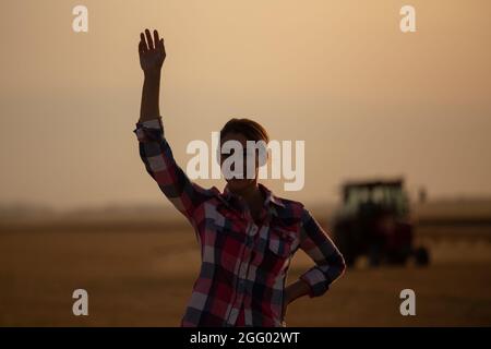 Une jolie jeune femme de campagne agite les mains devant le tracteur dans le champ au coucher du soleil Banque D'Images