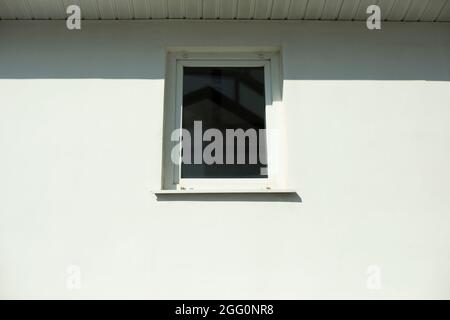 Fenêtre blanche dans un mur blanc. Une petite fenêtre en plastique aux rayons du soleil. Extérieur du bâtiment. Façade de mur blanchi. Banque D'Images