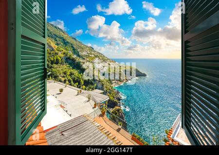 Vue à travers une fenêtre ouverte avec des volets de l'océan face à la colline ville de Praiano, Italie, le long de la côte amalfitaine. Banque D'Images