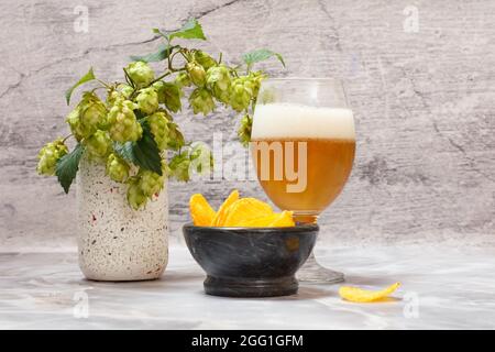 Verre de bière avec une branche de houblon et un bol avec des chips de pomme de terre sur fond gris. Banque D'Images