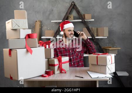 Le père Noël, propriétaire d'une petite entreprise, parle au téléphone sourit et prend une commande tout en étant assis à la table parmi les boîtes avec des cadeaux et des achats d'achat Banque D'Images