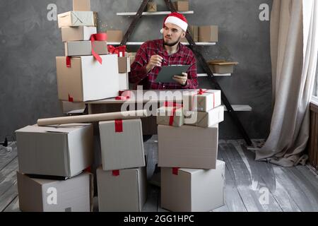 Un homme dans un chapeau de Père Noël travaille à la maison dans un entrepôt avec des boîtes de marchandises ou de cadeaux pour les enfants pour Noël. Comptabilité de marchandises magasin en ligne petit bus Banque D'Images