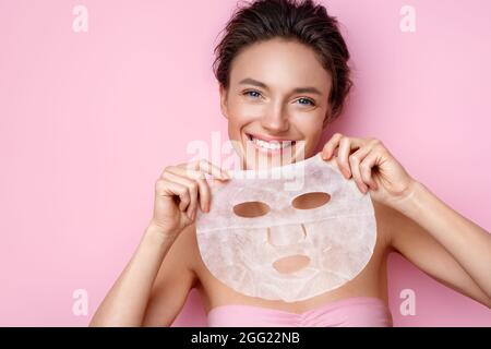 Une jeune femme souriante tient un masque cosmétique pour le visage. Photo d'une femme attrayante avec un maquillage parfait sur fond rose. Concept beauté et soin de la peau Banque D'Images