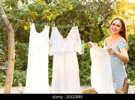 Jeune femme qui pendait du linge à l'extérieur. Mignonne fille en robe laver les vêtements blancs dans le lavabo en métal dans l'arrière-cour, pendant la lessive sur la corde à linge et de quitter Banque D'Images