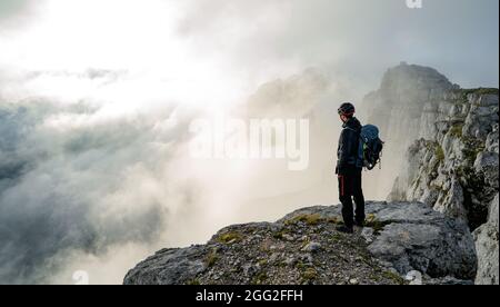 Homme avec sac à dos haut au-dessus de la vallée de montagne brumeux. La personne seule regarde une montagne entourée de brume et de nuages avec le pic visible. SC Banque D'Images