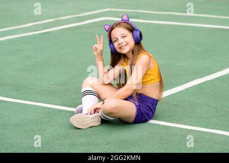 Chic et sympa, jeune fille d'école souriante, dans un casque sans fil, écoutant de la musique, portant des vêtements de sport tendance, se rafraîchissant sur un terrain de sport. Affiche de paix pour fille. Style boxer urbain Banque D'Images