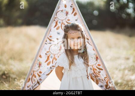 Joli mignon petit enfant fille modèle, portant élégant robe blanche et accessoires de plumes cheveux, regardant l'appareil photo, debout près de la tente de teepee dans le champ. Prise de vue en extérieur, style amérindien Banque D'Images