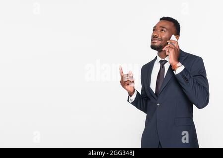 joyeux homme d'affaires afro-américain en costume ayant une conversation téléphonique tout en pointant vers le haut avec le doigt isolé sur le gris Banque D'Images