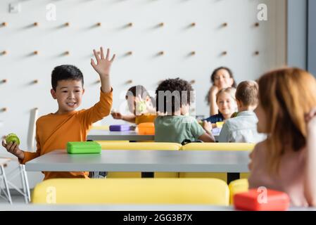 joyeux garçon asiatique en agitant la main à une fille floue dans le restaurant de l'école Banque D'Images