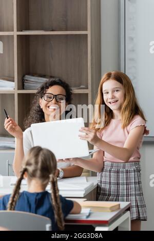 une jeune fille joyeuse tenant un ordinateur portable près d'un bon professeur afro-américain en classe Banque D'Images