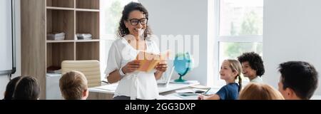 joyeux enseignant afro-américain avec livre souriant près des enfants multiethniques, bannière Banque D'Images