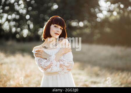 Portrait en plein air de jeunes femmes de style boho avec des tatouages flash sur le corps, portant une robe blanche debout dans le champ d'été et touchant sensuellement ses épaules. Concept de style amérindien Banque D'Images