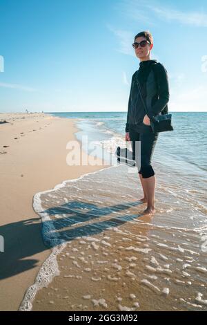 Photo verticale d'une femme dans des vêtements d'extérieur sombres avec des chaussures dans sa main droite, debout pieds nus sur une plage, avec la marée se lavant ses pieds. Magnifique Banque D'Images