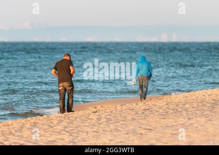 Deux hommes marchant sur une plage au bord de la mer Baltique loin du spectateur au début de la matinée d'été, avec le rivage dans la brume à l'horizon lointain. Après la fête. Banque D'Images