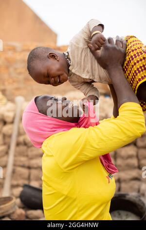 Une jeune mère portant un hijab islamique lève sa fille et lui sourit à l'extérieur de son domicile à Nouna, au Burkina Faso, en Afrique de l'Ouest. Banque D'Images