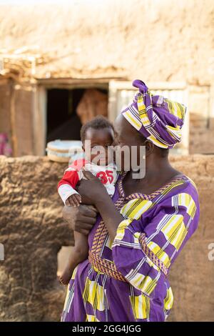 Mahammoud Traore (75, non représenté) soutient une famille de 21 personnes grâce à son agriculture dans le village de Dougouninkoro, Mali. Mais le changement climatique a affecté les tendances météorologiques ces dernières années et il n'a pas été capable de croître autant qu'il l'avait fait auparavant. Aujourd’hui, les réserves alimentaires de sa famille sont toujours à court de nourriture avant de pouvoir récolter sa nouvelle récolte. Par conséquent, il y a toujours deux mois chaque année où la famille fait l’expérience de la faim et qu’ils doivent souvent s’endetter pour acheter ou emprunter de la nourriture. Ici, la belle-fille de M. Traore, Oumou Dabou (36), tient sa fille Fatimata (9 mois), en dehors de la famille h Banque D'Images