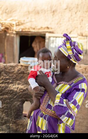 Mahammoud Traore (75, non représenté) soutient une famille de 21 personnes grâce à son agriculture dans le village de Dougouninkoro, Mali. Mais le changement climatique a affecté les tendances météorologiques ces dernières années et il n'a pas été capable de croître autant qu'il l'avait fait auparavant. Aujourd’hui, les réserves alimentaires de sa famille sont toujours à court de nourriture avant de pouvoir récolter sa nouvelle récolte. Par conséquent, il y a toujours deux mois chaque année où la famille fait l’expérience de la faim et qu’ils doivent souvent s’endetter pour acheter ou emprunter de la nourriture. Ici, la belle-fille de M. Traore, Oumou Dabou (36), tient sa fille Fatimata (9 mois), en dehors de la famille h Banque D'Images