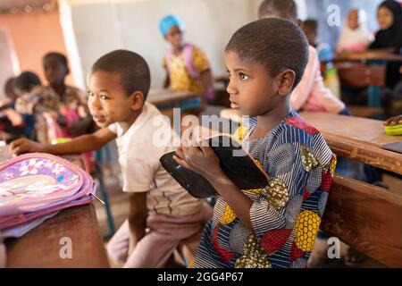 Le camp de Senou se trouve à la limite sud de Bamako, la capitale du Mali. C'est l'un des huit camps informels autour de la ville et, en soi, il abrite 223 familles et un peu plus de 1 000 personnes déplacées. Les familles qui vivent ici ont fui la violence et l'insurrection dans le nord et le centre du pays. Parce que la plupart d'entre eux n'ont pas de moyens de revenu ou de source régulière de nourriture, la faim est un défi quotidien et la malnutrition de sa population reste endémique. Ici, les enfants écrivent sur des lattes de craie dans la seule école primaire du camp, enseignée par Mme Asmatou Coulibably. Banque D'Images