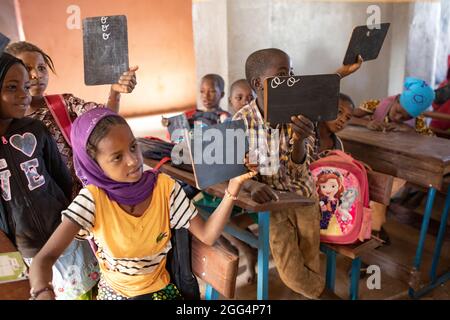 Le camp de Senou se trouve à la limite sud de Bamako, la capitale du Mali. C'est l'un des huit camps informels autour de la ville et, en soi, il abrite 223 familles et un peu plus de 1 000 personnes déplacées. Les familles qui vivent ici ont fui la violence et l'insurrection dans le nord et le centre du pays. Parce que la plupart d'entre eux n'ont pas de moyens de revenu ou de source régulière de nourriture, la faim est un défi quotidien et la malnutrition de sa population reste endémique. Ici, les enfants écrivent sur des lattes de craie dans la seule école primaire du camp, enseignée par Mme Asmatou Coulibably. Banque D'Images