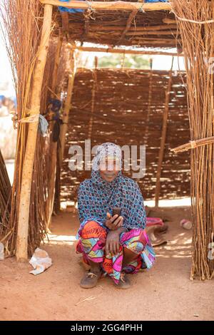 Le camp de Senou se trouve à la limite sud de Bamako, la capitale du Mali. C'est l'un des huit camps informels autour de la ville et, en soi, il abrite 223 familles et un peu plus de 1 000 personnes déplacées. Les familles qui vivent ici ont fui la violence et l'insurrection dans le nord et le centre du pays. Parce que la plupart d'entre eux n'ont pas de moyens de revenu ou de source régulière de nourriture, la faim est un défi quotidien et la malnutrition de sa population reste endémique. Ici, une femme est assise dans son abri de fortune tenant son téléphone portable. Banque D'Images