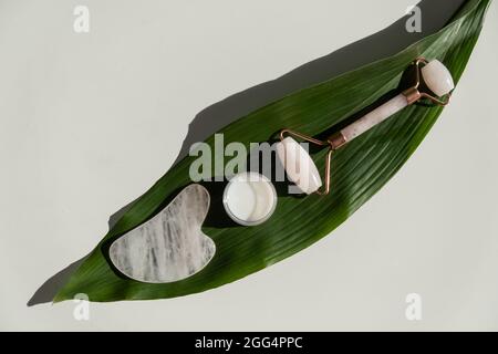 Grattoir de gouache rose, rouleau, pot de crème de feuille verte sur une table blanche. Équipement pour l'auto-massage et les soins de la peau pour le visage et le cou Banque D'Images