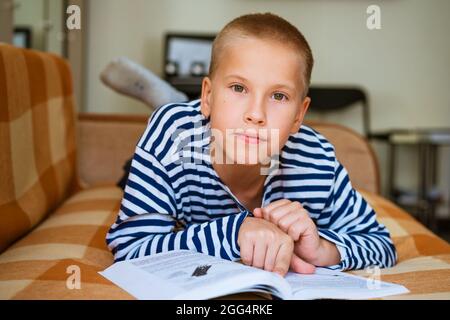 Un adorable garçon de dix ans qui fait ses devoirs à la maison sur un canapé allongé sur le dos pendant une pandémie ou après l'école. Mise au point sélective. Gros plan. Portrait d'un garçon d'école du caucase. Passe du temps utile à loisir Banque D'Images