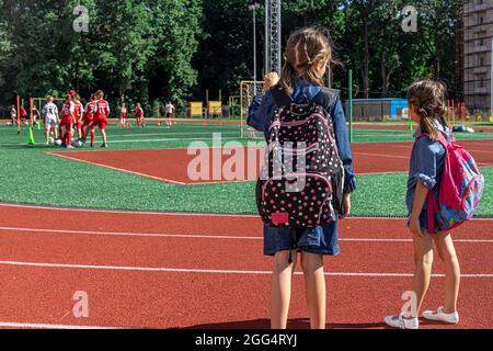 Les petites filles écoliers avec des sacs à dos dans le stade, en regardant les garçons jouer au football. Banque D'Images