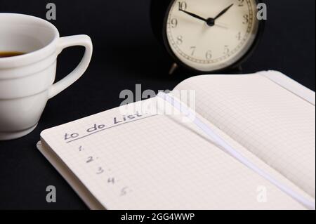 Image rognée d'un organiseur ouvert, bloc-notes avec liste à faire sur des feuilles blanches vierges, tasse de café, réveil sur fond noir avec espace de copie. Banque D'Images