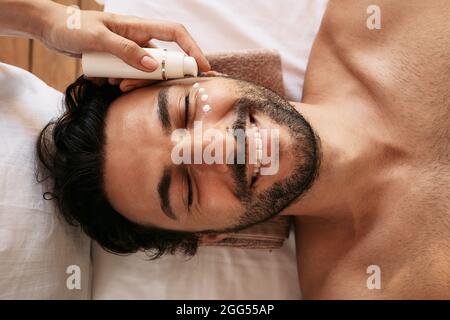 Une esthéticienne applique une crème hydratante pour la peau blanche au visage de l'homme au centre de bien-être. Le patient masculin bénéficie de soins de beauté, de soins de la peau et de détente à Banque D'Images