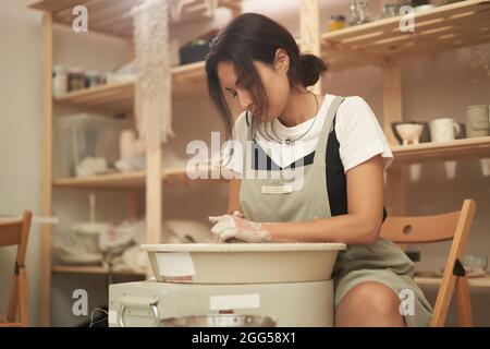 Jeune céramiste féminine en tablier, assise à la roue de poterie et à la modélisation du produit en argile tout en travaillant dans un studio d'art professionnel Banque D'Images