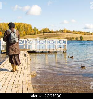 Une femme dans une veste et des bottes, avec un sac à dos, marche le long d'une jetée en bois dans un parc naturel et admire les oiseaux. Canards dans l'eau autour. Banque D'Images