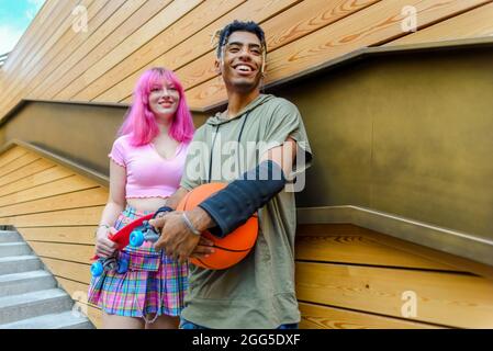 autre couple divers qui se traînait ensemble, se tenant debout contre le mur d'arrière-plan et souriant. heureux amis interraciaux tenant le skateboard Banque D'Images