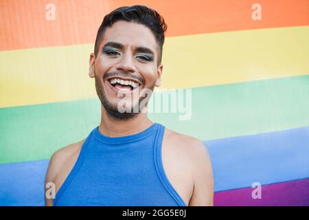 Jeune homme transgenre avec maquillage souriant sur la caméra avec le drapeau arc-en-ciel lgbt sur fond - Focus sur le visage Banque D'Images