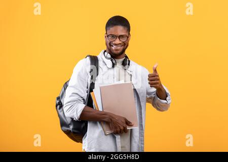 Étudiant afro-américain montrant le pouce vers le haut, posant avec sac à dos, ordinateurs portables et écouteurs sur le cou, fond jaune Banque D'Images