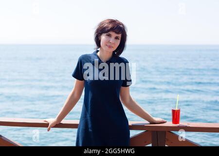 Une brunette souriante avec un verre de jus se tient sur la jetée au bord de la mer. Banque D'Images