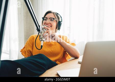 Une femme joyeuse hôte diffuse son podcast à l'aide d'un microphone vocal. Une femme qui fait du podcaster un flux audio dans son petit studio Banque D'Images