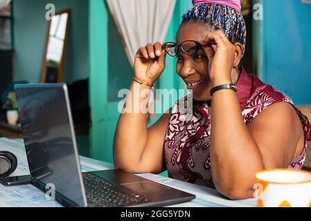 belle femme adulte africaine assis dans des vêtements légers porte des lunettes pour la vue devant elle, ayant travaillé devant un ordinateur pendant que smi Banque D'Images