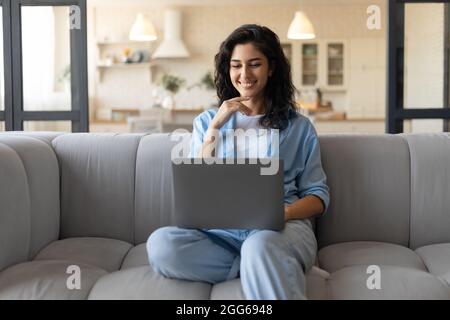 Travail indépendant, en ligne ou études. Jeune femme joyeuse assise sur un canapé avec ordinateur portable, travaillant ou apprenant de la maison Banque D'Images