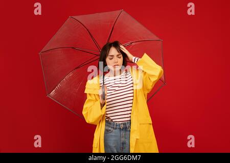 Femme millénaire tenant un parapluie transparent et touchant la tête, souffrant de maux de tête sur le mur rouge du studio Banque D'Images